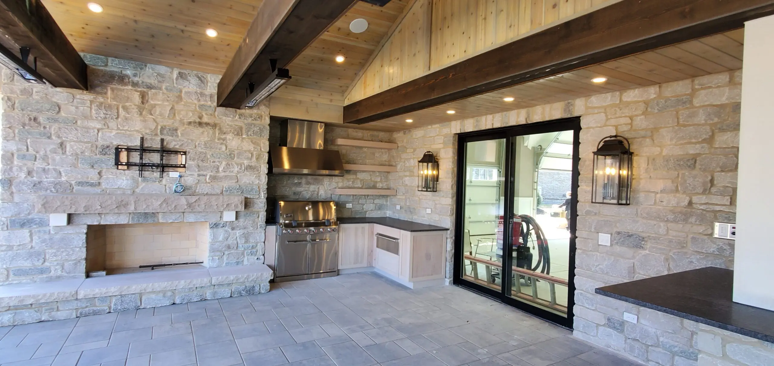 Modern outdoor kitchen with stone walls and built-in grill under wooden beams.